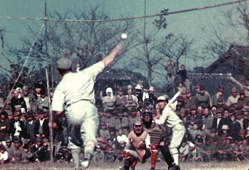 A Marine baseball player pitching to Japanese baseball player. Image shows three rows of people watching behind the mound.